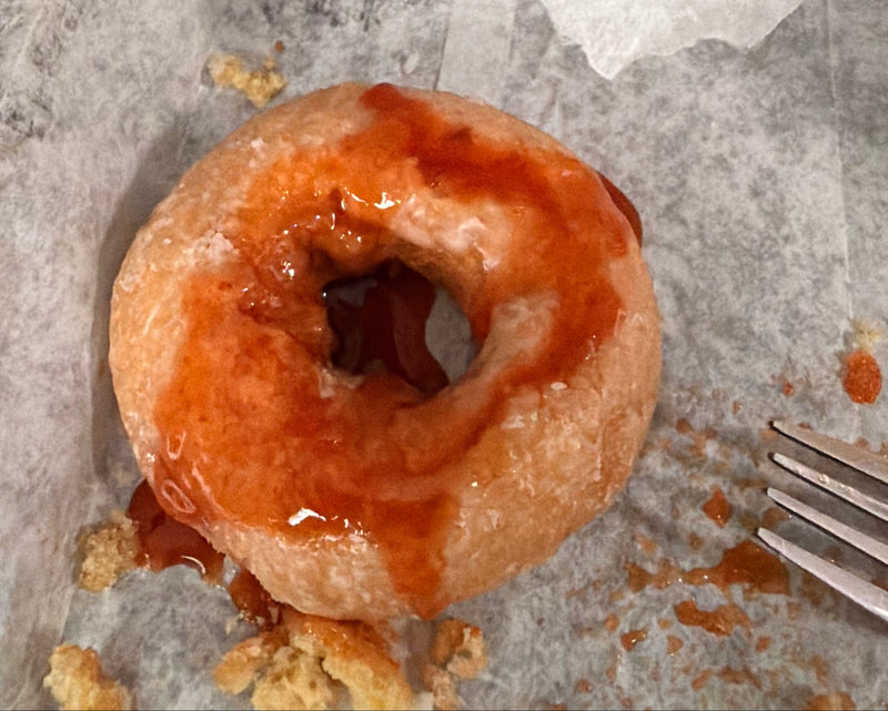 A glazed old fashioned donut with Nigel's Maple Fire poured on top  sits on torn white paper with crumbs and a fork visible.